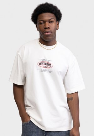 Unge mand med afro iført hvid oversize T-shirt med teksten "Off The Grid", guldkæde, blå jeans og tatovering på venstre underarm, poserer.