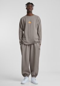 Langermet grå skjorte med oransje logo og komfortable grå joggebukser med elastiske mansjetter, kombinert med hvite sneakers på en ensfarget bakgrunn.