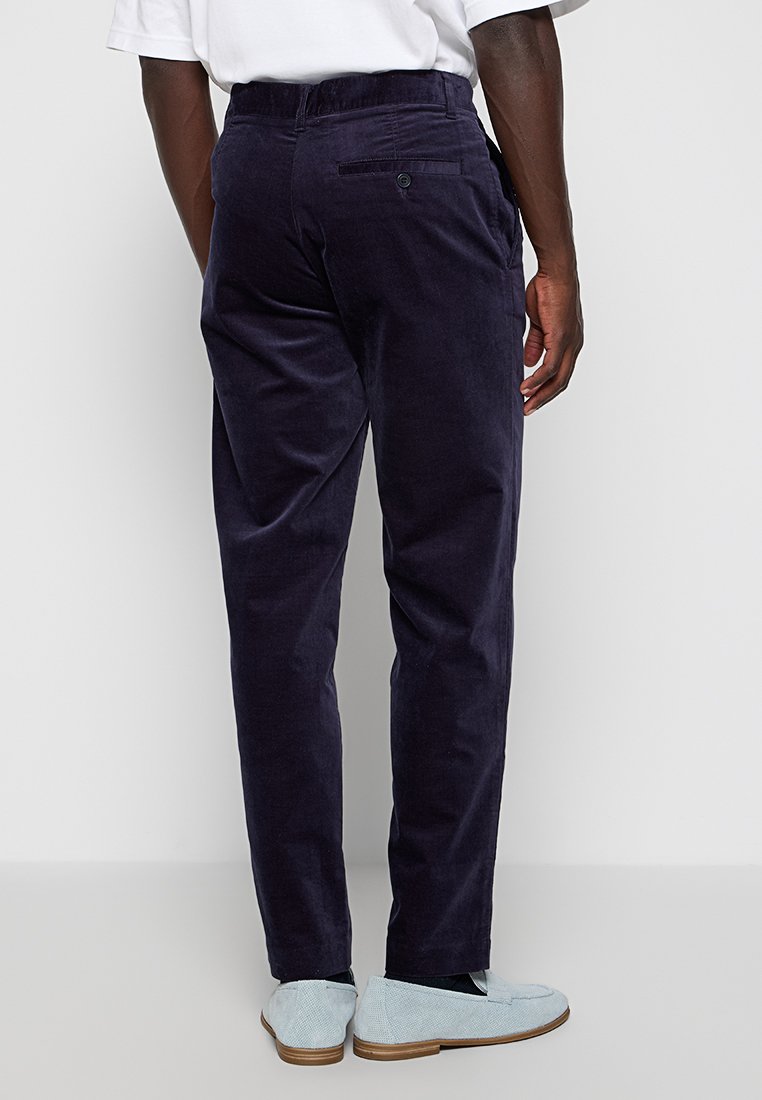 Man in een donkerblauwe corduroy broek, wit shirt gestoken, en lichtblauwe instappers, staand tegen een effen lichte achtergrond.