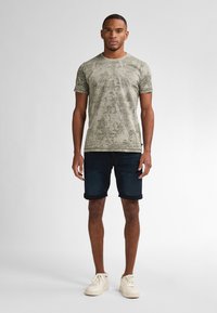 Vaate lyhythihaisesta, vaaleasta oliivinvihreästä t-paidasta, jossa on tekstuuri kuvio, yhdistettynä tummansinisistä farkkushortseista ja vaaleista lenkkareista.