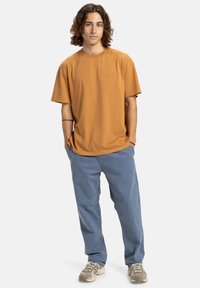Jonge man met krullend haar, gekleed in een mosterdkleurig t-shirt, blauwe broek en beige sneakers, staand met zijn handen in de zakken tegen een witte achtergrond.