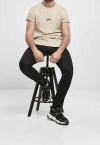 Muž v béžovém tričku "OFF.", černých kalhotách a černo-bílých teniskách sedící na černém nastavitelném stoličce na bílém pozadí.
