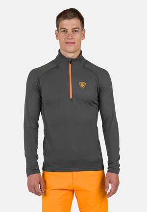 Camicetta a maniche lunghe con zip di colore grigio scuro con design testurizzato, accentuata da dettagli arancioni e logo del marchio sul petto. Abbinata a pantaloni arancioni brillanti.