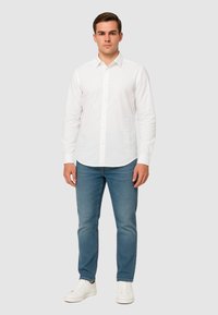 Camicia bianca button-up in tessuto leggero con maniche lunghe, abbinata a jeans blu e sneakers bianche. Dettaglio testuale sul lato della camicia.
