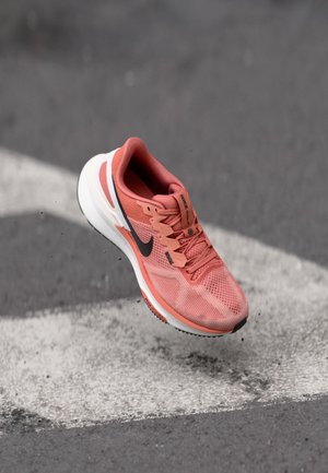 Zapatilla de running en malla color coral con acentos negros, con una suela blanca y superficie texturizada, suspendida sobre un fondo gris con una franja.
