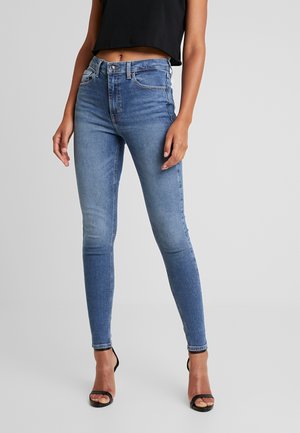 Niebieskie jeansy w kroju slim fit, noszone z czarnym krótkim topem oraz czarnymi sandałami na obcasie z odkrytymi palcami, stojące na jednolitym jasnoszarym tle.