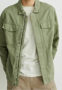 Světle zelená bunda vyrobená z měkké tkaniny, s límcem, předními patentkami a dvěma náprsními kapsami. Nositá přes bílou košili.