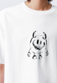 Tricou alb din bumbac cu un grafic negru al unui personaj rotund cu coarne și o față zâmbitoare, cu mâneci scurte și guler rotund.