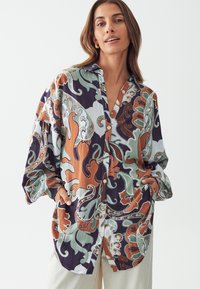 Camisa de manga larga con botones que presenta un patrón floral abstracto multicolor, principalmente en tonos de azul marino, verde, naranja y blanco, con un corte relajado.