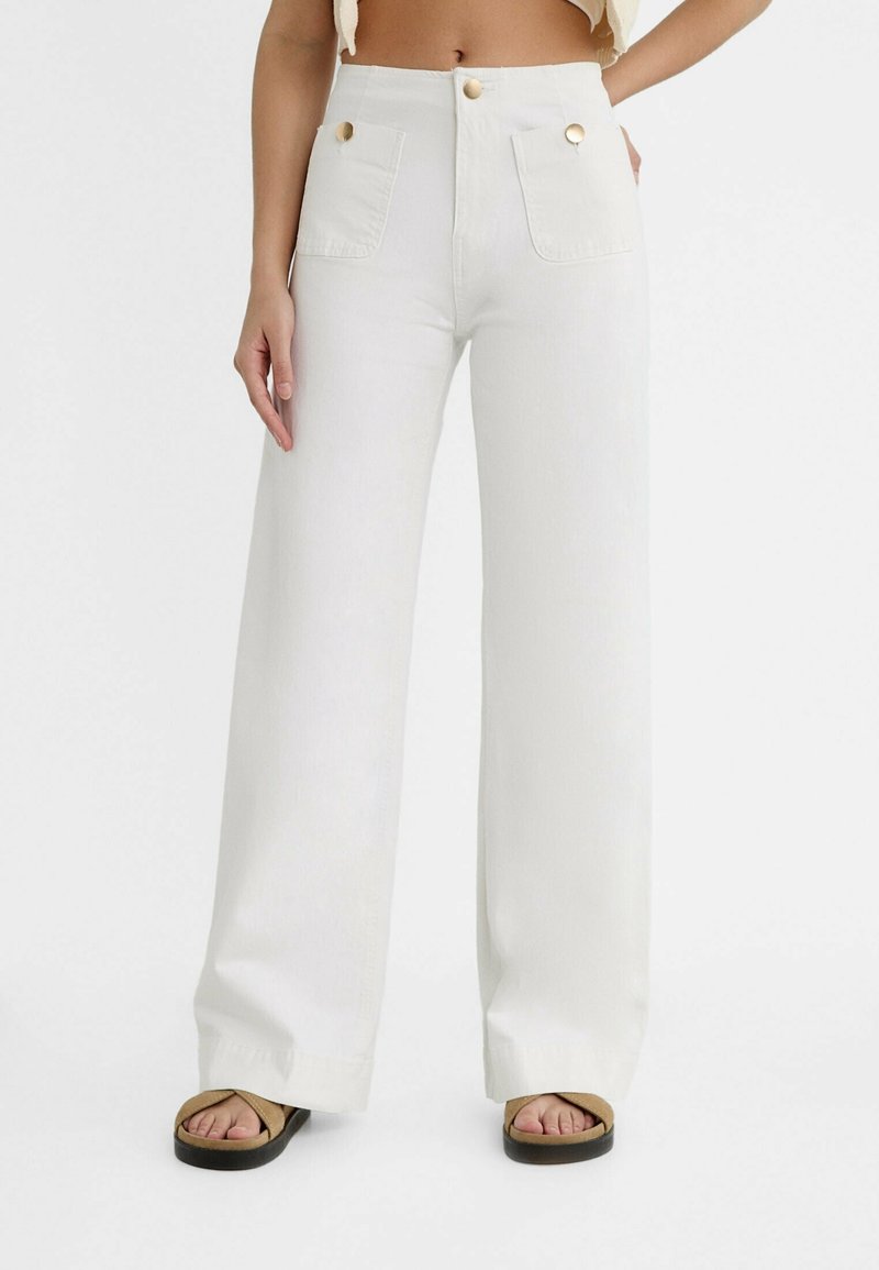 Femme portant un pantalon blanc taille haute à jambes larges avec poches avant et boutons dorés, associé à des sandales couleur sable sur fond blanc.
