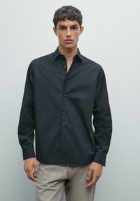Giovane uomo con capelli corti e scuri, indossa una camicia nera con bottoni e pantaloni grigio chiaro, in piedi davanti a uno sfondo grigio semplice.