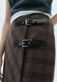 Jupe à carreaux bruns avec un design superposé, dotée de sangles en cuir noir et d'accents en matériel argenté. Texture douce et coupe ajustée.