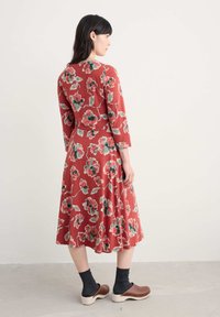 Femeie în picioare de profil, purtând o rochie florală roșie, șosete închise la culoare și saboți din lemn maro, pe un fundal neutru simplu.