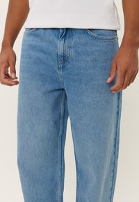 Light blue denim jeans with a relaxed fit, visible stitching details, and a smooth texture. Worn with a white ribbed shirt.