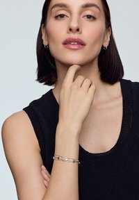 Srebrna bransoletka z kwiatowymi akcentami, spleciony wzór, noszona na nadgarstku; na neutralnym tle z ciemnym bezrękawnikem.