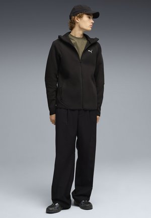 Jonge persoon met een zwarte pet, een zwarte hoodie met rits en Puma-logo, een olijfgroen shirt, losse zwarte broek en zwarte sneakers, staand en naar rechts kijkend.