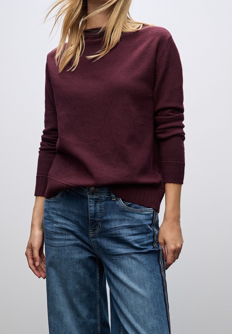 Femme portant un pull à manches longues bordeaux et un jean bleu avec une bande latérale, debout devant un fond gris clair uni.