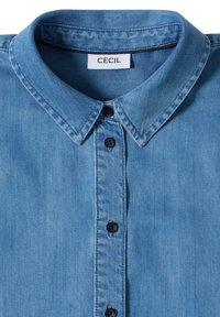 Chemise en denim bleu clair avec un col pointu et trois boutons foncés visibles, avec une étiquette blanche portant le texte « CECIL » à l’intérieur du col.