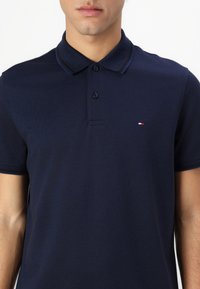 Námornícky modrá polo košeľa vyrobená z textúrovaného materiálu s trojica gombíkov a kontrastným golierom. Malé logo v červenej, bielej a modrej farbe.