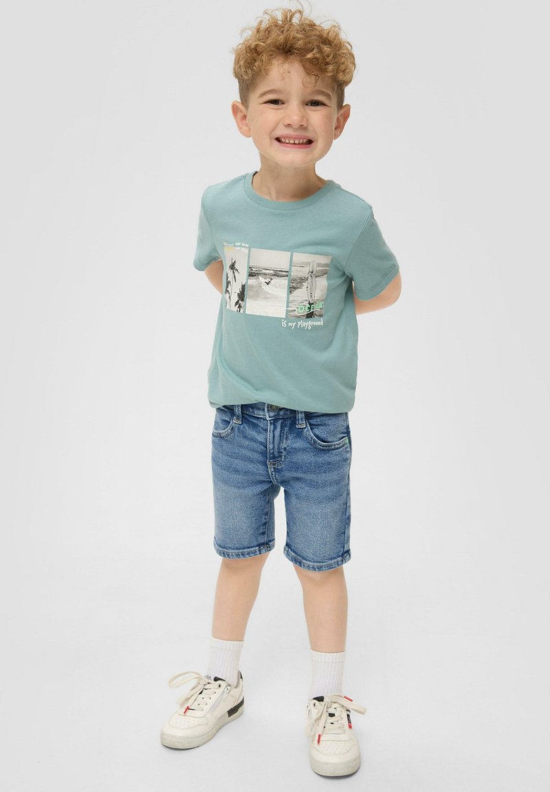 Jasno-niebieska bawełniana koszulka z nadrukami zdjęć, zestawiona z dżinsowymi szortami. Białe trampki z szarymi akcentami i żebrowane białe skarpetki dopełniają całość stroju.