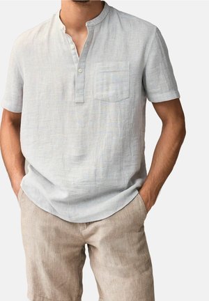 Mand iført lys grå henley t-shirt med korte ærmer og brystlomme samt beige linnedshorts, med hænderne i lommerne, foran en ensfarvet baggrund.