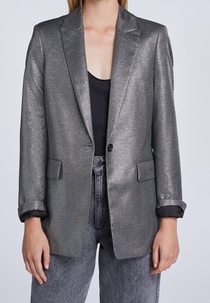 Frau trägt ein strukturiertes, metallisch silbernes Sakko über einem schwarzen Top und grauen hoch taillierten Jeans, steht vor einem schlichten hellen Hintergrund.