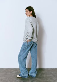 Mujer de pie de lado, vistiendo un sudadera gris, jeans azules holgados y zapatillas blancas, con un fondo de pared blanca lisa.