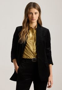 Blazer di velluto nero con una camicia di raso dorato sotto. L'outfit include una cintura con una fibbia d'argento ornata e pantaloni neri attillati.