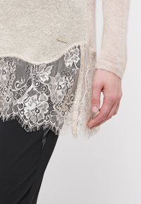 Pull en maille beige avec détail d'ourlet en dentelle florale et petite étiquette métallique "Guess", porté avec un pantalon noir sur le bas du torse d'une personne.