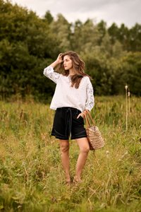 Femme en blouse blanche brodée et short noir debout dans un champ herbeux, tenant un sac fourre-tout tissé, avec des arbres en arrière-plan.