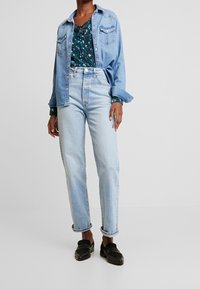 Jasnoniebieskie dżinsy o prostym kroju, w zestawieniu z ciemną bluzką w wzory i jasnoniebieską koszulą zapinaną na guziki. Czarne buty typu slip-on.