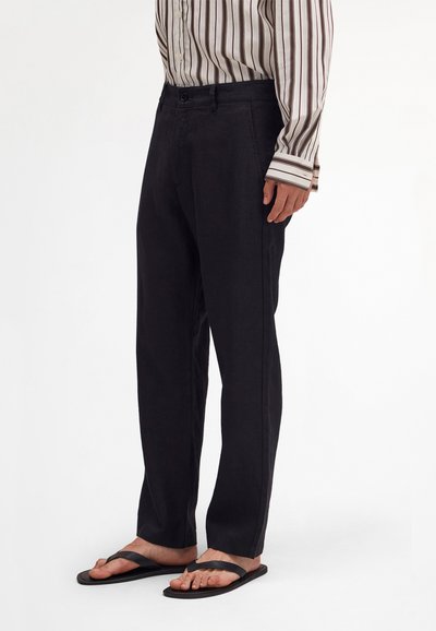 Mann trägt schwarze, gerade geschnittene Hosen, ein gestreiftes langärmliges Hemd, das in die Hose gesteckt ist, und schwarze Flip-Flops. Er steht vor einem weißen Hintergrund.