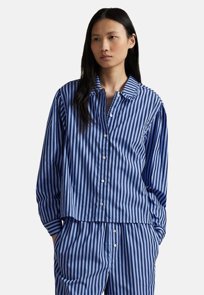 Femme portant une chemise boutonnée à rayures verticales bleues et bleu foncé ainsi qu'un pantalon assorti, les mains dans les poches, expression neutre.