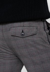 Grå rutiga byxor med röda accenter, som har en bakficka med knappstängning och en slät textur. Midjebandet är svart.