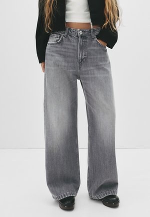 Person iført højtnærede, vidde-ben grå denimjeans, sorte sko, sort jakke og hvid croppet top mod en ensartet lys baggrund.