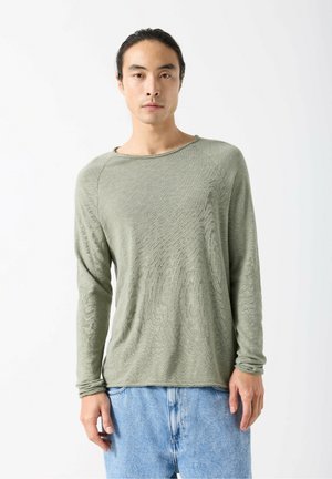 Pikkade varrukatega, oliivroheline kampsun tekstuurse pinnaga, ümar kaelus ja raglaanvarrukad, ühendatud helesiniste teksadega.