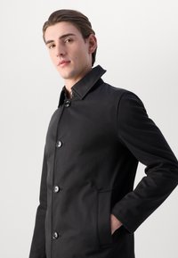 Giovane uomo con pelle chiara e capelli castani, vestito con un cappotto nero bottonato con tasche frontali, in piedi contro uno sfondo chiaro e semplice.