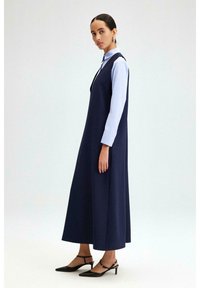Robe sans manches bleu marine avec un col montant, portée sur une chemise à manches longues bleu clair, associée à des escarpins noirs à bout pointu. Tissu lisse.