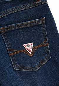 Jeans in denim blu con un distintivo patch triangolare rosso e bianco sulla tasca posteriore, accentuato da cuciture arancioni a contrasto.