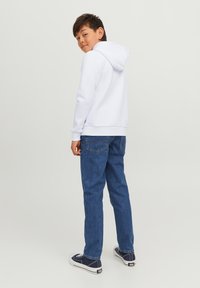 Felpa bianca con cappuccio, dalla vestibilità comoda, abbinata a jeans blu di denim e sneakers in tela. Il ragazzo è leggermente girato, mostrando l'outfit.