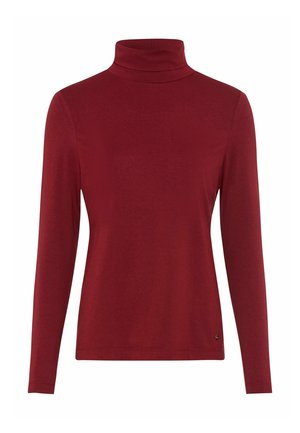 Kvinde iført en bordeaux langærmet top, beige bukser, hvide sneakers, et cremefarvet tørklæde og holder en ternet frakke over skulderen.