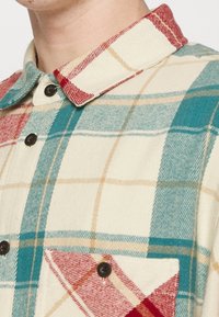 Rutet skjorte i creme, teal og rød med knapkrave. Har en brystlomme og sorte knapper. Blødt, tekstureret stof.