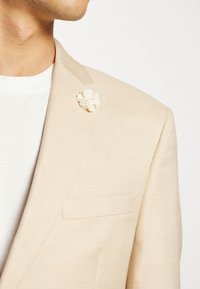 Sacou bej cu un material texturat, având un singur buzunar pe piept și o broșă florală albă, complicată, pe rever.