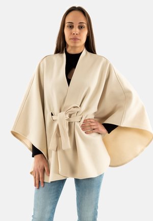 Donna che indossa un mantello beige con cintura sopra una maglia nera e jeans blu, in piedi con una mano sui fianchi contro uno sfondo bianco uniforme.