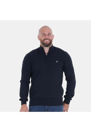 Homme chauve souriant portant un pull bleu marine foncé avec un col à fermeture éclair et un petit logo sur la poitrine, debout devant un fond uni.