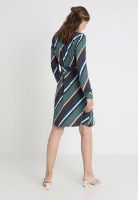 Vihreä mekko, jossa on pitkät hihat, esittelee vinoraitoja tummansinisessä, vaaleansinisessä ja oranssissa. Mukana on solmittava vyö vyötäröllä ja keskipitkä leikkaus.