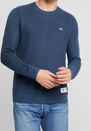 Homme portant un pull en maille bleu marine texturé avec des manches et un ourlet côtelés, associé à un jean bleu clair, les mains lâchement jointes.