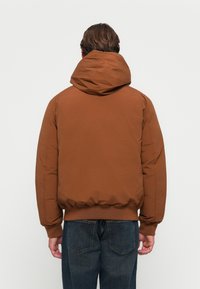 Brązowa pikowana kurtka z kapturem, elastycznymi mankietami i pasem. Gładka tekstura z minimalnymi detalami przeszycia, noszona na ciemnych dżinsach.