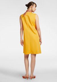 Abito giallo senza maniche con taglio dritto, caratterizzato da una sciarpa stampata al collo e tacchi aperti arancioni. Tessuto dalla texture liscia.