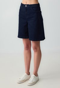 Pantaloni shorts in denim blu navy con taglio dritto, cinque tasche e chiusura con bottone. Indossati con sneakers bianche, caratterizzate da una superficie testurizzata.